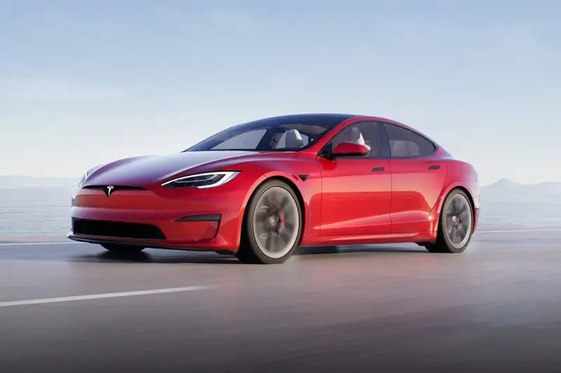 Une Tesla Model S Plaid rouge vif file sur une route côtière, le soleil se reflétant sur sa carrosserie élégante.