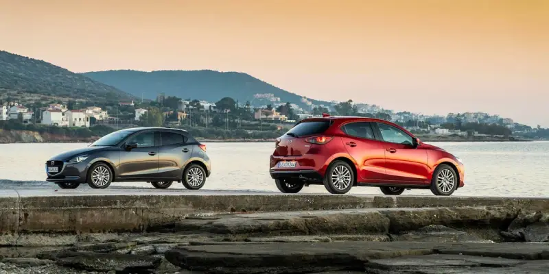 Dos Mazda, uno gris y otro rojo, junto al mar. La fiabilidad Mazda se refleja en su diseño y presencia.