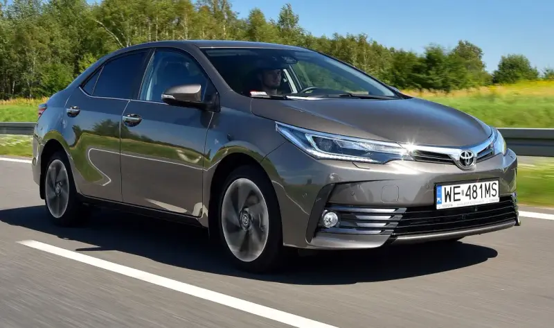 Szara Toyota Corolla E16 jedzie po drodze, za kierownicą siedzi mężczyzna.