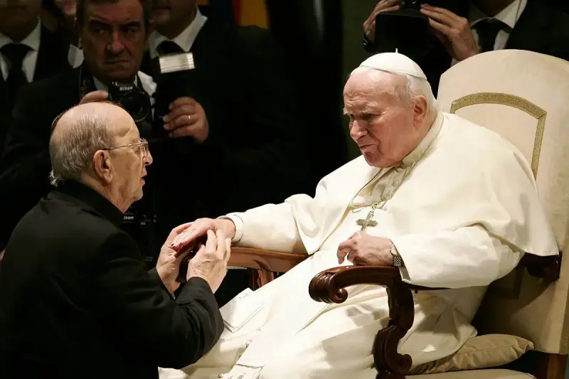 Papież Jan Paweł II w białej szacie, klęczący przed nim mężczyzna w czerni. W tle widać innych duchownych i dziennikarzy, być może z zakonu **jezuitów**.
