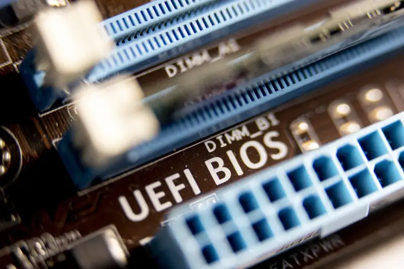 Zbliżenie na płytę główną z widocznym napisem "UEFI BIOS" i złączami RAM. W tle widać oznaczenie DIMM_A1, a poniżej DIMM_B1. Ten układ jest kluczowy dla bezpieczeństwa systemu, podobnie jak moduł **TPM 2.0**.