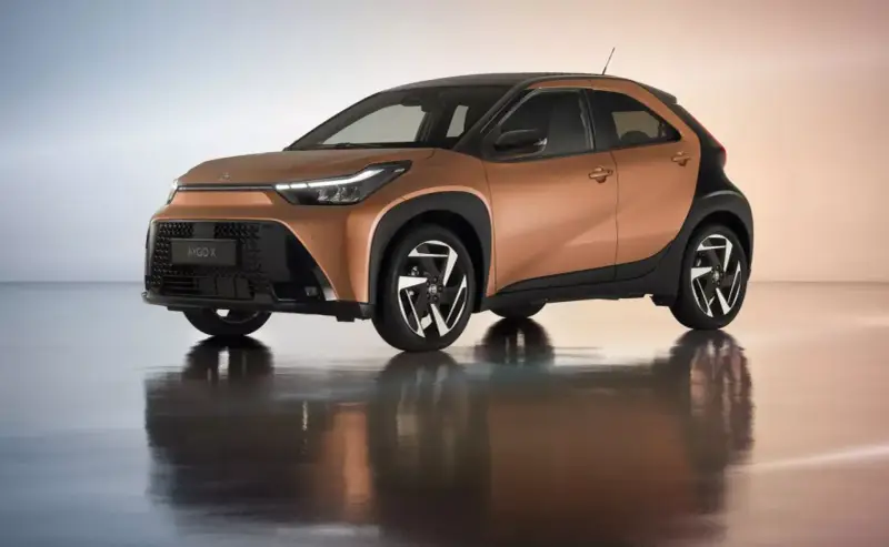 Brązowy Toyota Aygo X z czarnym dachem i felgami na błyszczącym podłożu.