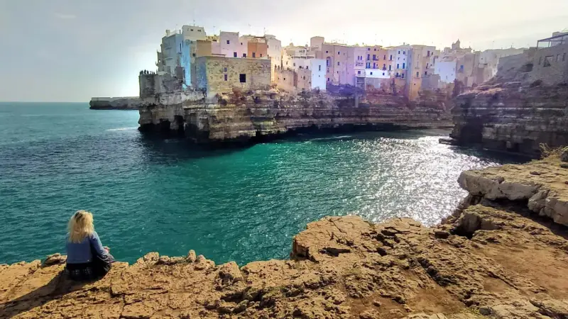 Kobieta siedzi na skale, podziwiając widok na Polignano a Mare. Białe domy wznoszą się na klifie nad turkusowym morzem.