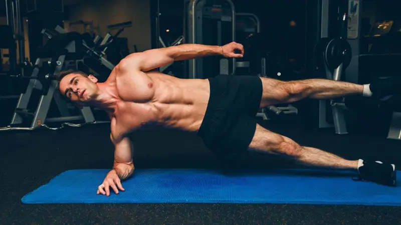 Un homme musclé réalise un gainage planche sur un tapis bleu dans une salle de sport.