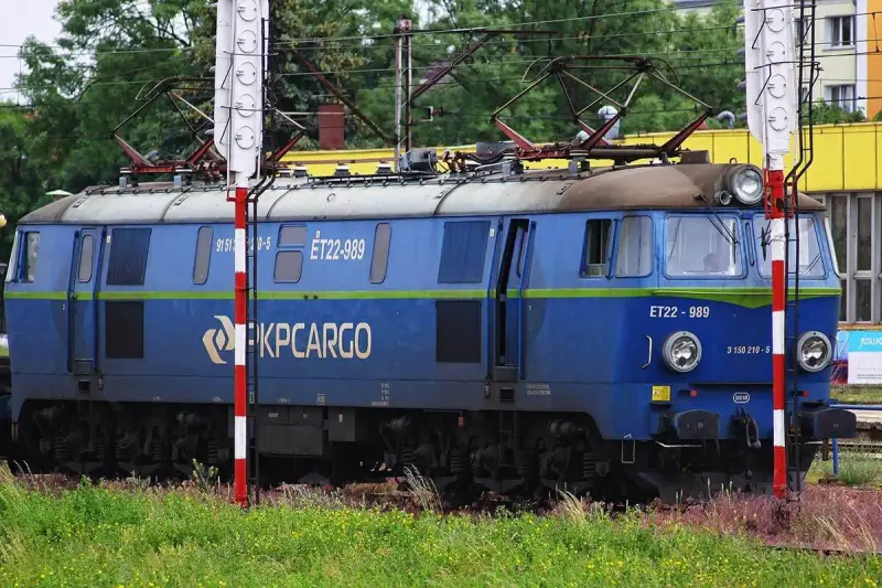 Niebieska lokomotywa PKP Cargo ET22-989 na tle zieleni i budynków.