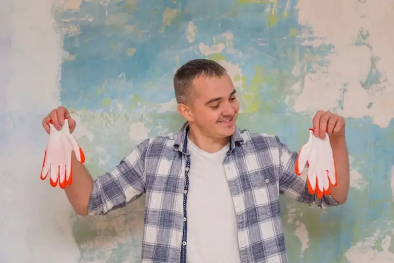 Homme souriant tenant des gants blancs à bouts orange devant un mur texturé. Il pense au prix peinture m2 au black pour son projet.