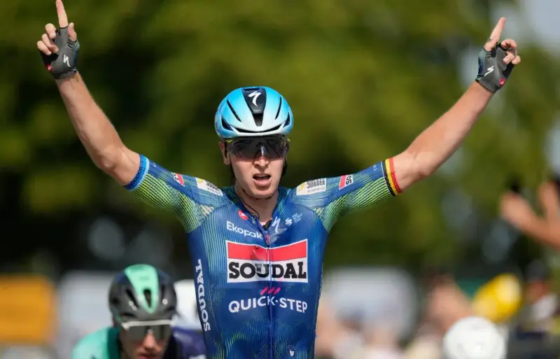 Tim Merlier jubelt mit erhobenen Armen und zeigt den Sieg. Er trägt ein blau-grünes Radtrikot mit Sponsorenlogos.