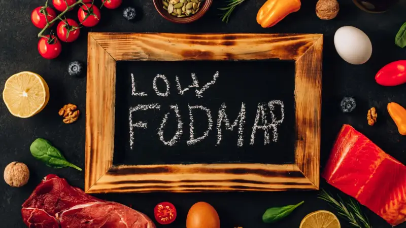 Na tablicy napis "LOW FODMAP". Wokół świeże produkty: mięso, ryba, pomidorki, cytryna, jajko, papryka, orzechy, jagody, szpinak.