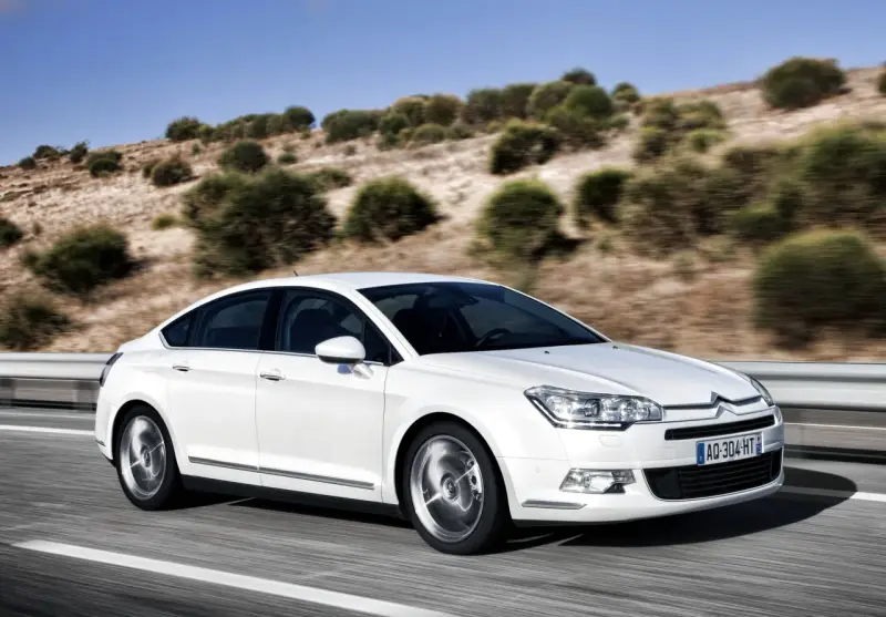 Biały Citroen C5 2.0 HDI pędzi po pustej drodze, otoczony suchymi, pagórkowatymi krajobrazami.