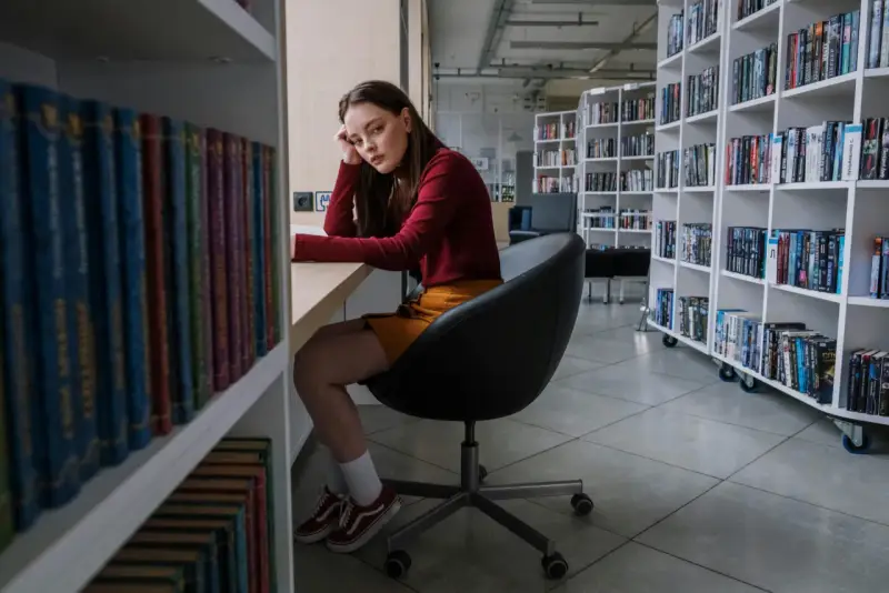 Zamyślona nastolatka w bibliotece, otoczona książkami, odzwierciedla złożoność zachowań nastolatków w okresie dojrzewania.