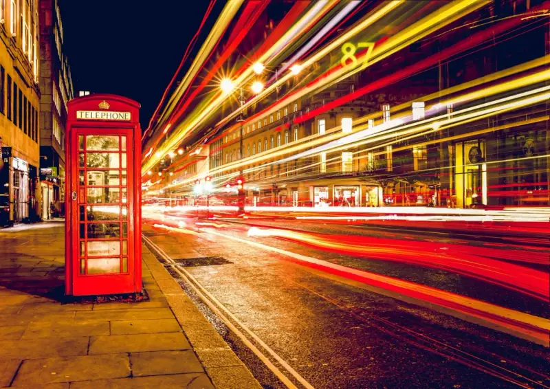 Klasyczna czerwona budka telefoniczna na tle nocnej ulicy, gdzie długi czas naswietlania zdjecia uchwycił smugi świateł samochodowych.