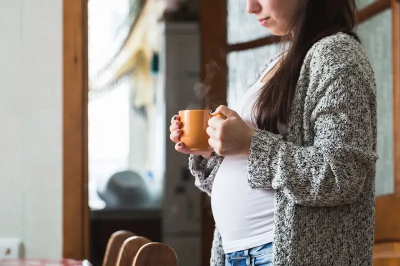 Ciepły napar z rooibos w dłoniach ciężarnej kobiety. Delikatny dym unosi się z kubka, tworząc chwilę spokoju.