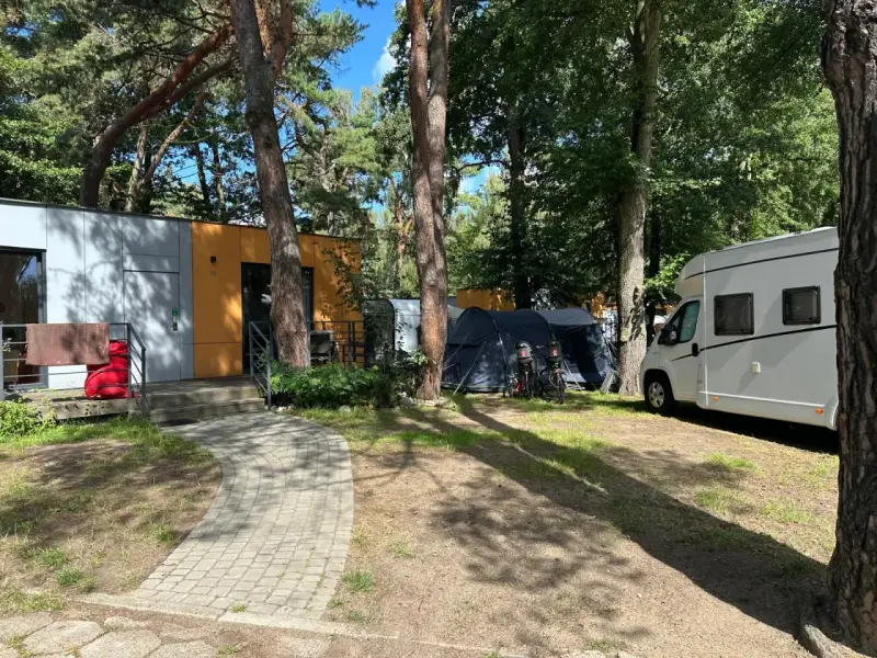 Na kempingu 45, wśród drzew, stoją nowoczesne domki, namioty i biała kamper.