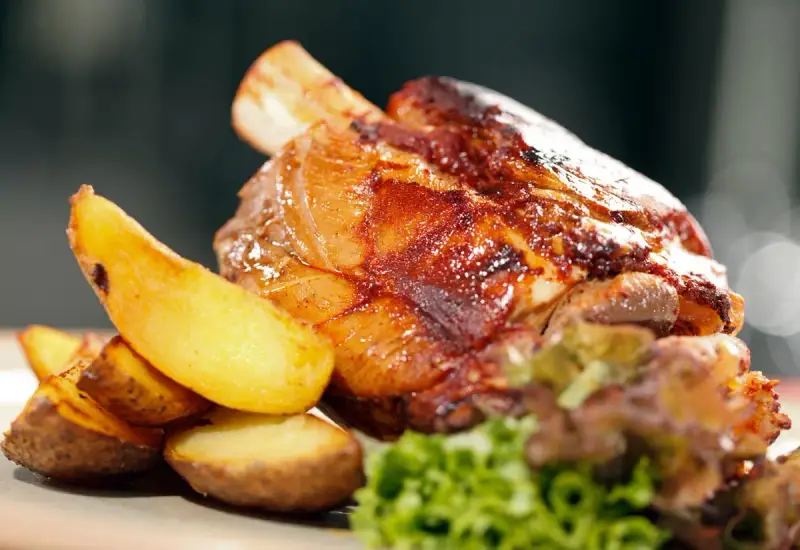 Codillo de cerdo asado, crujiente y jugoso, acompañado de patatas gajo doradas y una guarnición verde.