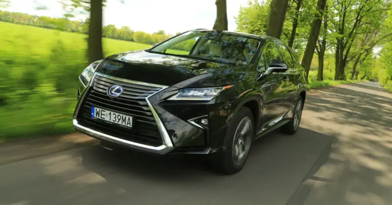 Czarny Lexus RX 450h pędzi po wiejskiej drodze, otoczony zielenią drzew i pól.