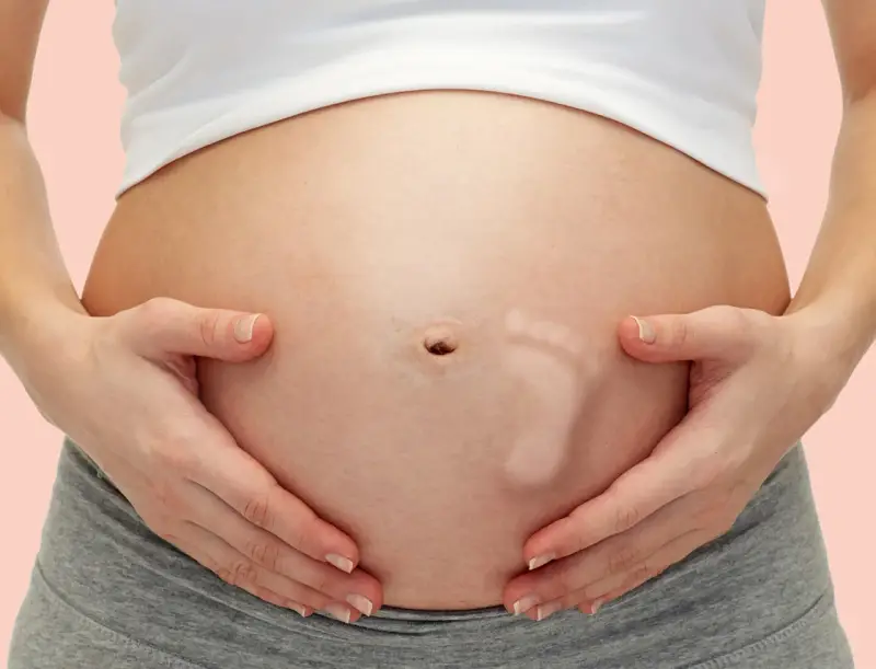Brzuszek w 30 tygodniu ciąży, z widocznym odciskiem stópki dziecka. Ręce mamy delikatnie obejmują rosnący brzuch.