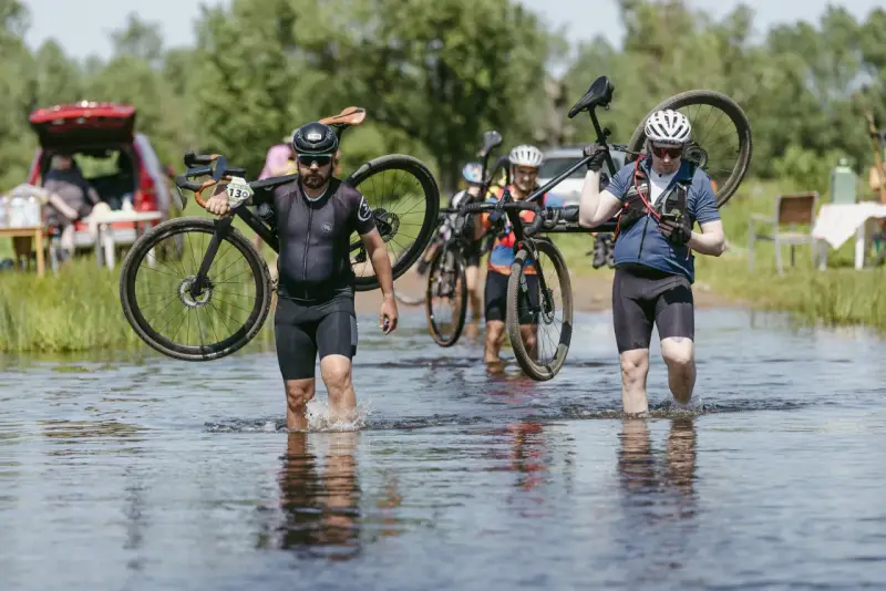 Kolarze pokonują wodną przeszkodę podczas mazowiecki gravel. Niosą rowery przez płytką rzekę, w tle zielone drzewa i samochód.