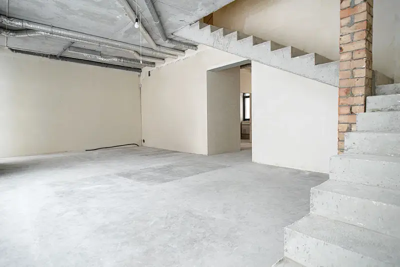 Wnętrze w stanie deweloperskim: betonowe schody, nieotynkowane ściany, widoczne instalacje i przewody.