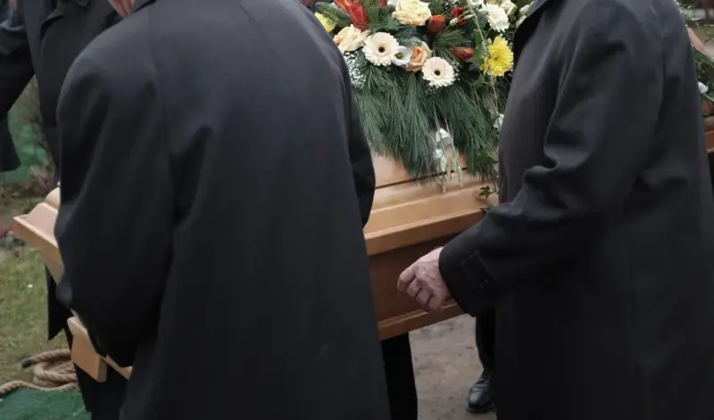 Mężczyźni w czarnych płaszczach niosą trumnę ozdobioną kwiatami. Kondukt pogrzebowy w żałobnej ciszy.
