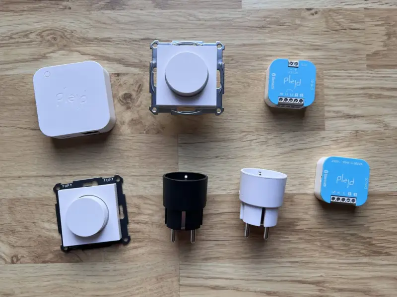En samling Plejd-produkter, inklusive en vit Plejd-box, två dimmerknappar, två elkontakter och två styrenheter som stöder Plejd-protokoll.