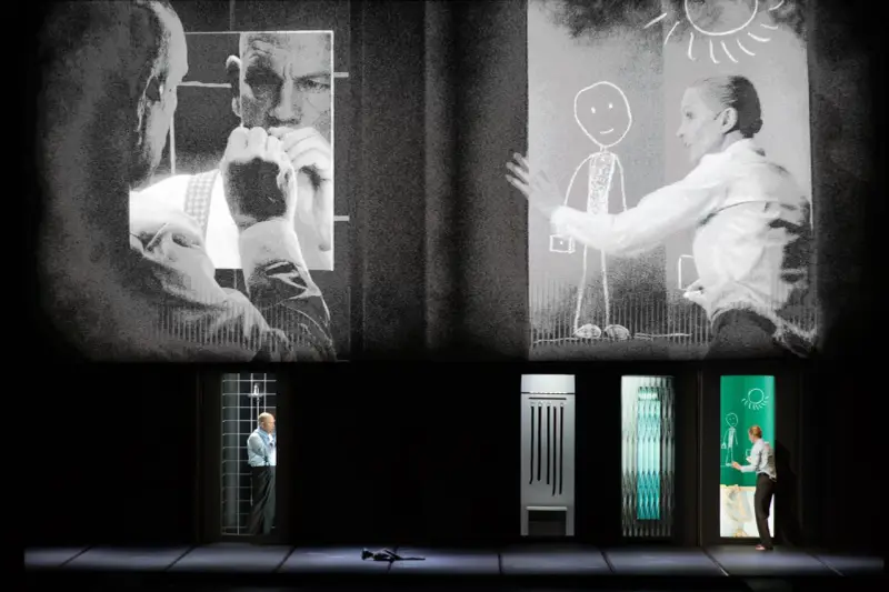 Scena teatralna z projekcjami. Na górze czarno-białe obrazy mężczyzn i kobiety rysującej słońce. Na dole, w oknach, samotność pól bawełnianych, mężczyzna w klatce, przedmioty i kobieta rysująca.