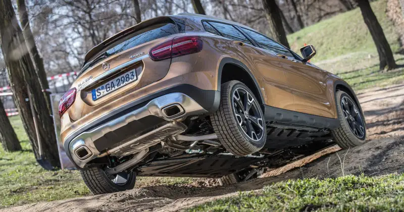 Złoty Mercedes GLA 220 4MATIC pokonuje nierówny teren, unosząc jedno koło.