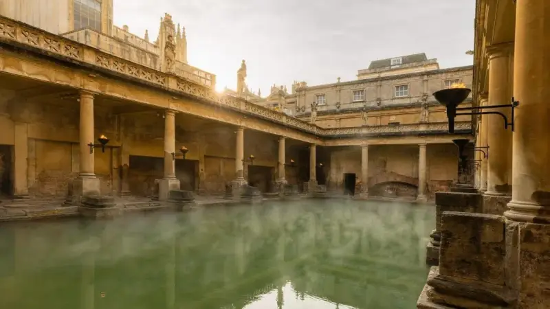 Steaming green water fills the Great Bath, surrounded by ancient Roman ruins with columns and torches.