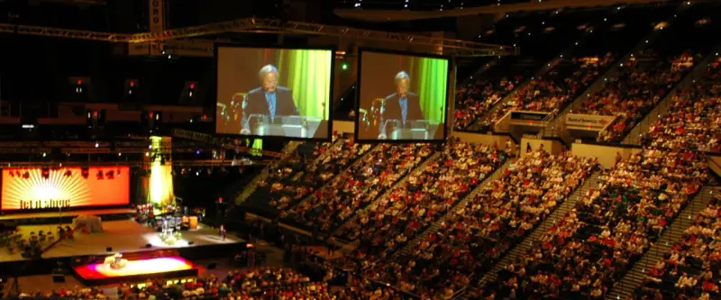 Große Menschenmenge in einer Arena, die einer Predigt von der **United Church of Christ** auf einer Leinwand folgt.