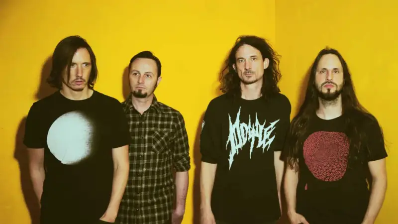 Quatre hommes aux cheveux longs posent devant un fond jaune vif. Leurs t-shirts portent des graphismes distinctifs, évoquant l'énergie brute du magma et la puissance de Gojira.