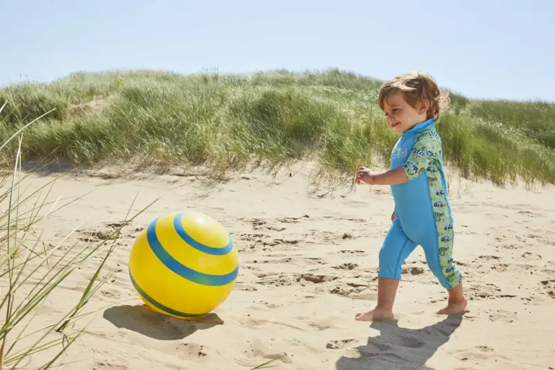 Chłopiec w niebieskim stroju kąpielowym z neoprenu bawi się żółtą piłką na plaży.