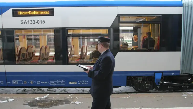 Konduktor w mundurze stoi przy pociągu i trzyma tablet. W oknie widać pasażerów.