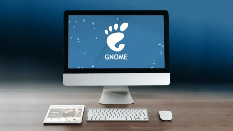 Komputer z logo GNOME, tablet i klawiatura na drewnianym biurku.