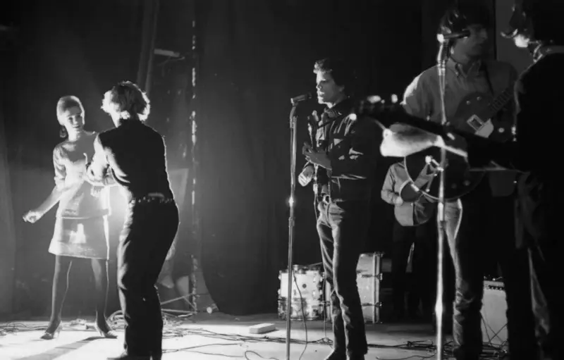 Scène de concert des membres de The Velvet Underground. Une femme danse à gauche, tandis que Lou Reed chante au micro.