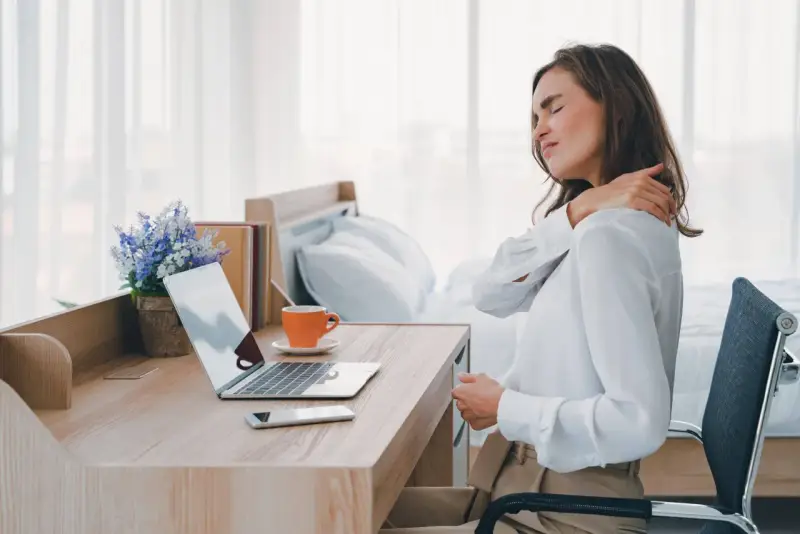 Femme se massant l'épaule, assise à son bureau. La mauvaise position bureau peut causer des douleurs.