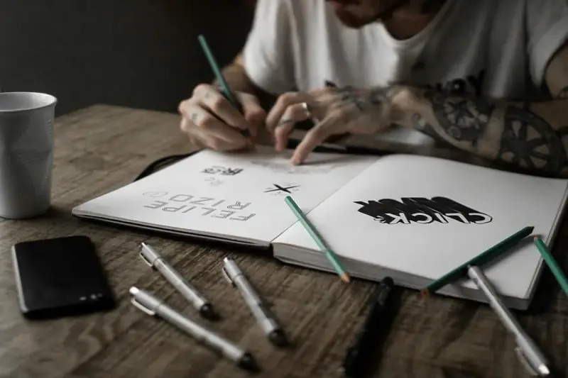 Un artiste auteur ou micro entrepreneur dessine dans son carnet, entouré de stylos, crayons et d'une tasse.