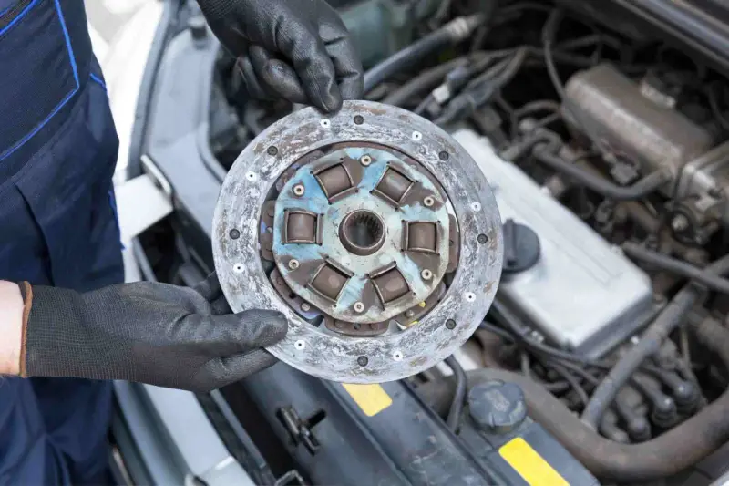 Mechanik w czarnych rękawiczkach trzyma zużyte sprzęgło samochodowe na tle silnika.