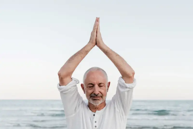 Mężczyzna z zamkniętymi oczami, ręce złożone nad głową, praktykuje mindfulness co to. Spokój nad morzem.