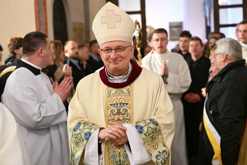 Uśmiechnięty biskup w stroju pontyfikalnym, otoczony przez wiernych i duchownych. W tle widać fragmenty kościoła, a na pierwszym planie szczegóły szat.