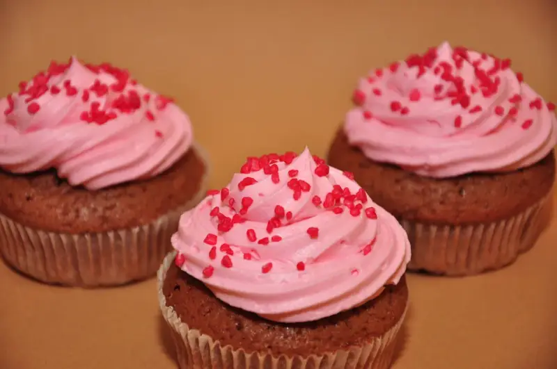 Tre chokladmuffins med rosa frosting och röda strössel, bakade i rosa muffinsformar.