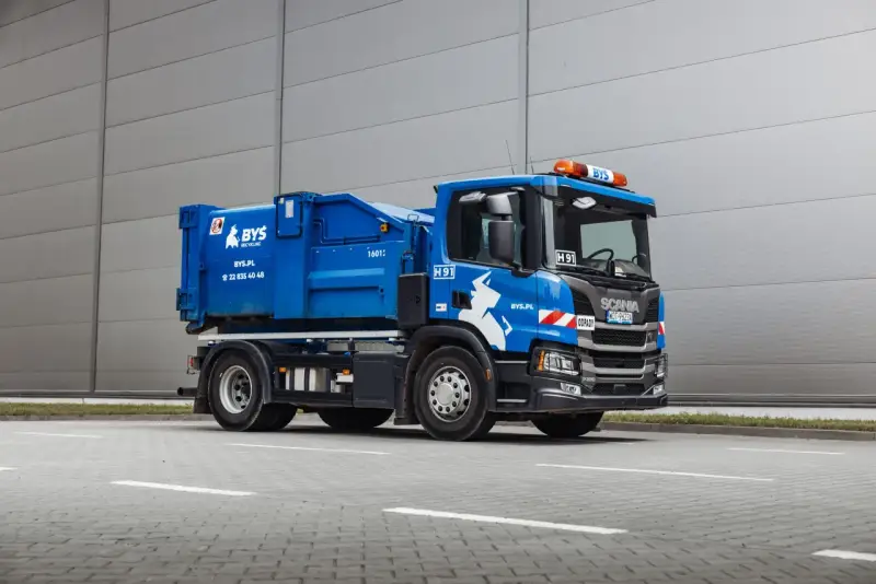 Niebieska śmieciarka Scania z napisem "ODPADY" i logo BYS. Pojazd gotowy do odbioru gruzu, z kodem odpadu na tablicy.