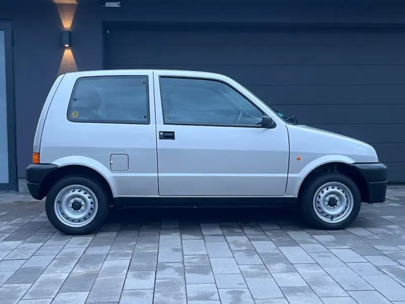 Srebrny Fiat Cinquecento na tle ciemnego garażu, z charakterystycznymi felgami i czarnymi zderzakami.