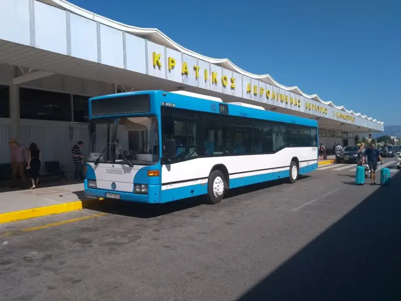 Niebiesko-biały autobus na tle budynku lotniska z żółtym napisem.