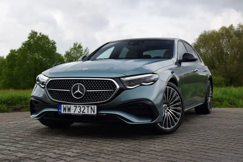 Elegancki Mercedes E220d w kolorze miętowym, zaparkowany na brukowanej nawierzchni, z zielenią w tle.