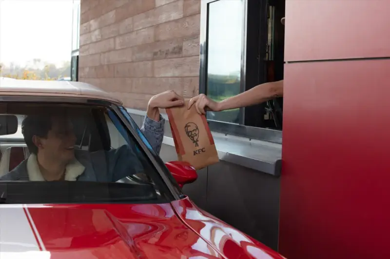 Mężczyzna w czerwonym aucie odbiera z okienka KFC torbę z jedzeniem. Wygląda na zadowolonego, że otrzymał swój mega pocket kcal.