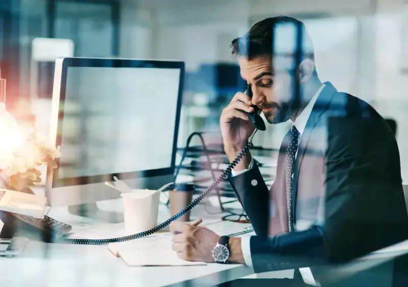 Biznesmen w garniturze rozmawia przez telefon, notując w zeszycie. Nawet w obliczu siły wyższej, praca wre.