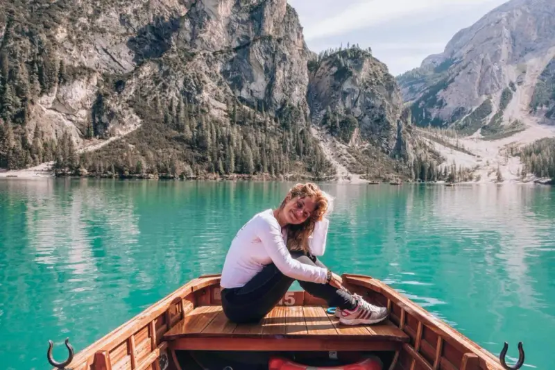 Uśmiechnięta kobieta w łodzi na turkusowym jeziorze w **Włoszech Dolomity**. Majestatyczne góry i lasy tworzą tło dla tej malowniczej sceny.