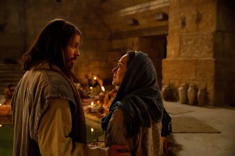 Scena z Kany Galilejskiej: Jezus rozmawia z kobietą, w tle widać gości weselnych i świece.