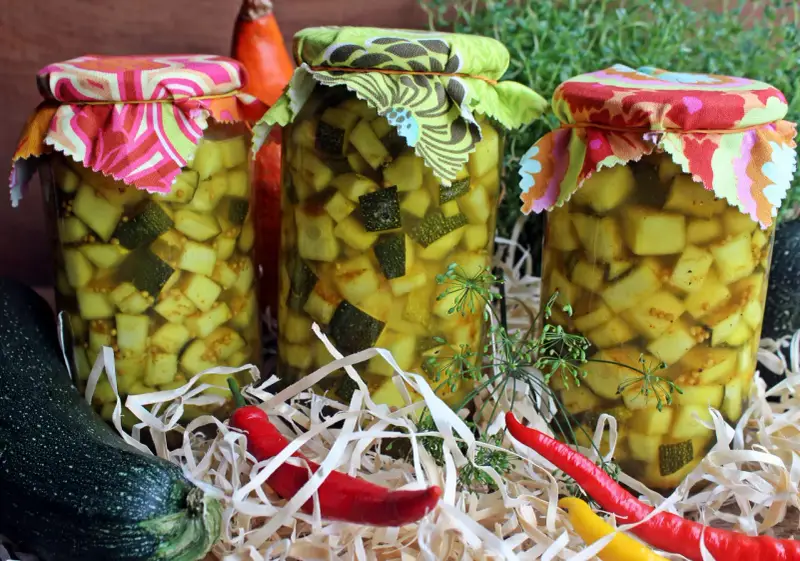 Słoiki z cukinią w zalewie słodko-kwaśnej z curry, ozdobione kolorowymi materiałami. Obok leżą świeże cukinie i papryczki chili.