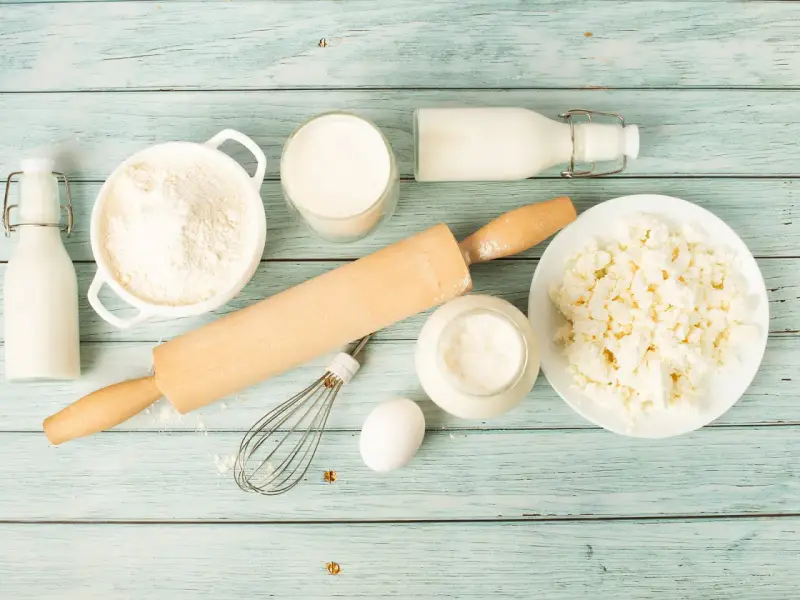 Zutaten für butter ersatz backen: Mehl, Milch, Quark, Ei, Schneebesen und Nudelholz auf blauem Holzuntergrund.