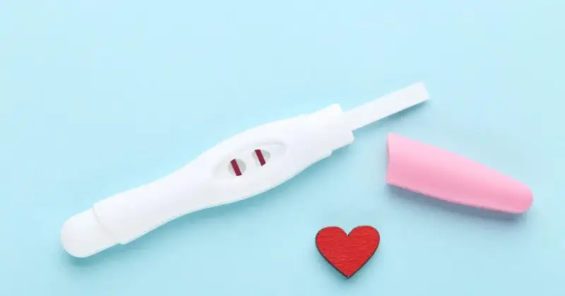 Biały test ciążowy z dwoma czerwonymi paskami, różowa nakładka i czerwone serduszko na niebieskim tle.